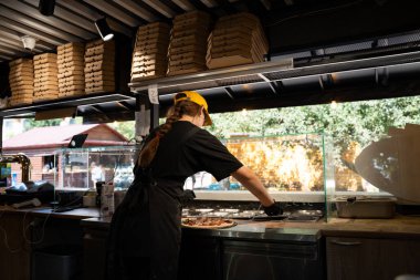 Şef, kalabalık bir büfede pizza hazırlıyor. Doğal ışık ve pizza kutuları üst üste yığılmış..