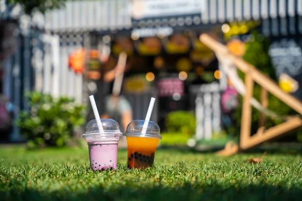 A lone cup of pink bubble tea with a straw, set against a blurred background of a vibrant street food stall.