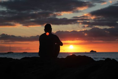 Man sitting alone on beach rocks looking at the sunset, lost in thought or contemplation