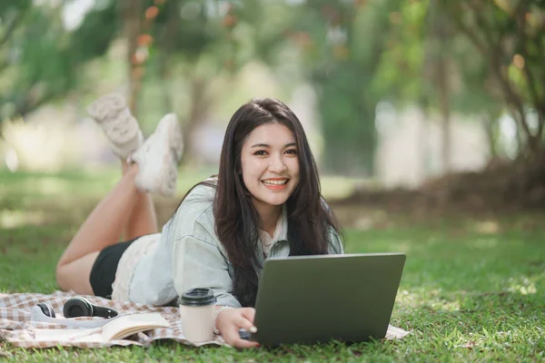 Asian woman lying on picnic blanket and lawn at park working on laptop. Asian female using laptop while sitting under a tree at park with bright sunlight. Work from anywhere concept.