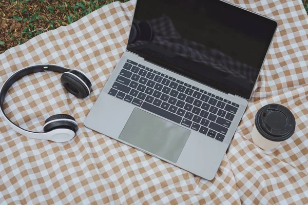Work on laptop on picnic blanket on the lawn in the park- next to a headphone, notebook  and cup of hot coffee. Freelancer work concept. Top view, flat lay. Work form anywhere concept.