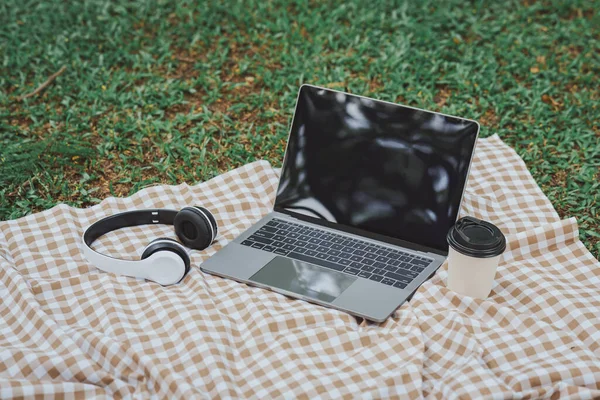 Work on laptop on picnic blanket on the lawn in the park- next to a headphone, notebook  and cup of hot coffee. Freelancer work concept. Top view, flat lay. Work form anywhere concept.