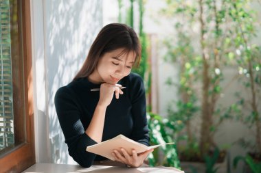 Siyah gömlek giyen Asyalı çalışan bir kadın ve kapalı kafedeki masanın üzerine küçük bir defter üzerine günlük yazıyor. Kadın notları ve kafede kahve içmesi. Herhangi bir yerden çalışma kavramı.