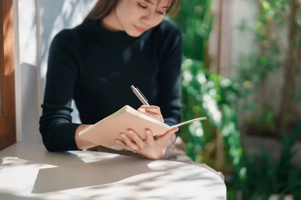 黒いシャツを着たアジア人の働く女性が屋内カフェのテーブルの上に小さなノートに日記を書いています カフェで女性のノートやコーヒーを飲む コンセプトはどこからでも — ストック写真