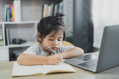 Mavi çizgili gömlek giyen Asyalı bir kız bebek evdeki oturma odasındaki ahşap masa masasında çalışmak için dizüstü bilgisayar kullanıyor ve not defteri ile notlar yazıyor. Eğitim öğrenimi online olarak ev kavramından.