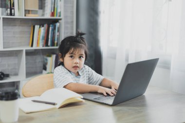 Mavi çizgili tişört giyen Asyalı bebek evdeki oturma odasındaki tahta masada çalışmak için dizüstü bilgisayar ve defter kullanıyor. Eğitim öğrenimi online olarak ev kavramından.