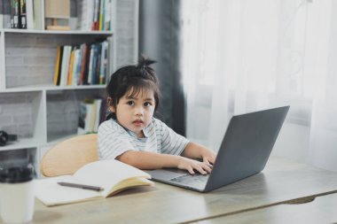 Mavi çizgili tişört giyen Asyalı bebek evdeki oturma odasındaki tahta masada çalışmak için dizüstü bilgisayar ve defter kullanıyor. Eğitim öğrenimi online olarak ev kavramından.