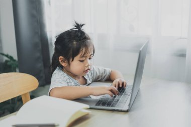 Mavi çizgili tişört giyen Asyalı bebek evdeki oturma odasındaki tahta masada çalışmak için dizüstü bilgisayar ve defter kullanıyor. Eğitim öğrenimi online olarak ev kavramından.