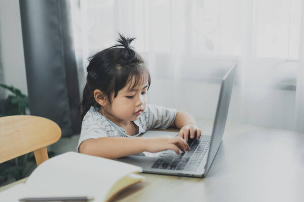 Asian baby girl wearing a blue striped shirt use laptop and notebook to study online on wood table desk in living room at home. Education learning online from home concept.
