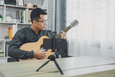 Asyalı müzik öğretmeni cep telefonuyla öğrencilere canlı gitar dersleri veriyor. Gitar çalan Asyalı erkek müzisyen evde canlı video konferansı yapıyor. Herhangi bir yerden canlı konser müziği.