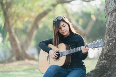 Kafasında uzun kollu siyah bir gömlek ve güneş gözlüğü olan Asyalı bayan müzisyenler. Sabahları parktaki bir ağacın altında akustik gitar çalıp şarkı söylemek..