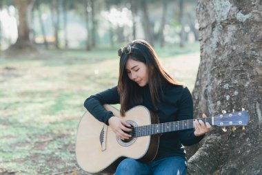 Kafasında uzun kollu siyah bir gömlek ve güneş gözlüğü olan Asyalı bayan müzisyenler. Sabahları parktaki bir ağacın altında akustik gitar çalıp şarkı söylemek..