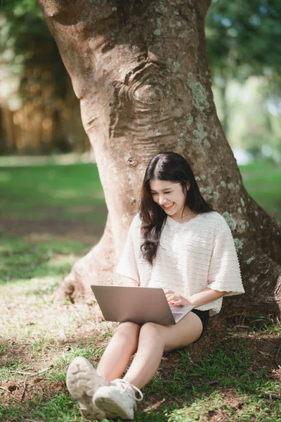 stock image Asian woman sitting on picnic and lawn at park working on laptop. Asian female using laptop while sitting under a tree at park with bright sunlight. Work from anywhere concept