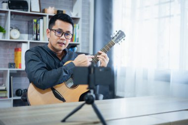 Asyalı müzik öğretmeni cep telefonuyla öğrencilere canlı gitar dersleri veriyor. Gitar çalan Asyalı erkek müzisyen evde canlı video konferansı yapıyor. Herhangi bir yerden canlı konser müziği.