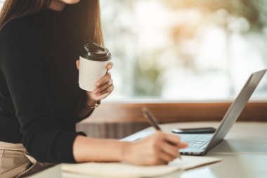 Asian freelance woman smiling holding cup of hot coffee and working on laptop computer on wooden table at home. Entrepreneur woman working for her business at home. Business work at home concept.