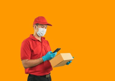 Cerrahi maske takan Asyalı kurye, sarı arka planda izole edilmiş kırmızı üniformalı tıbbi eldiven, mobil kontrol stokları kullanarak ve posta yoluyla paket göndermek veya taşımak için paket kutuları tutmak
