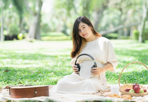 Pregnant happy asian woman putting headphones on her belly to listen music while sitting on picnic blanket on lawn at the garden park under the tree. Family mother mom pregnant healthy concept.