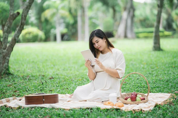 Pregnant happy asian woman using tablet to chat or social media or browse internet while sitting on picnic blanket on lawn at garden park under the tree. Family mother mom pregnant healthy concept.