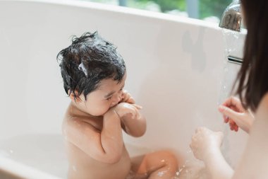 Asyalı tatlı kız çocuğu annesiyle banyo yaparken saçlarını yıkıyor banyo küvetini temizliyor. Anne sevgisi ve ilgisiyle mutlu bebek masum suyun tadını çıkarırken iyi eğlenceler. Bebek konsepti.