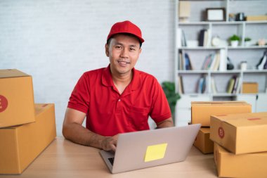 Asian delivery man worker smiling in red uniform work at home , checking list parcel boxes for sending or conveying parcels by mail. Delivery transport concept.
