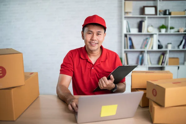 Asian delivery man worker smiling in red uniform work at home , checking list parcel boxes for sending or conveying parcels by mail. Delivery transport concept.