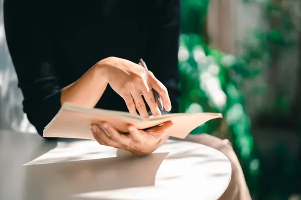 stock image working woman wearing black shirt and writing journal on small notebook on the table at indoor cafe. Woman notes and drinking coffee at cafe. Working from anywhere concept.