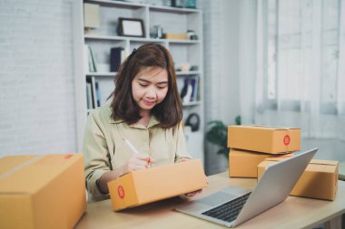 Asyalı kurye kadın evde çalışıyor, paket kutularını posta yoluyla gönderme veya taşıma için işaretliyor.