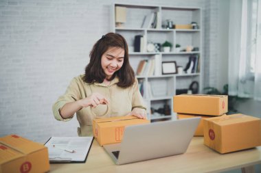 Asian delivery woman freelance smiling and packing box of products working at home , checking list parcel boxes for sending or conveying parcels by mail.