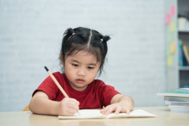 Kırmızı tişörtlü Asyalı bir kız bebek evdeki oturma odasındaki tahta masada ders çalışmak için deftere notlar yazar ve kitap okur. Eğitim öğrenimi online olarak ev kavramından.