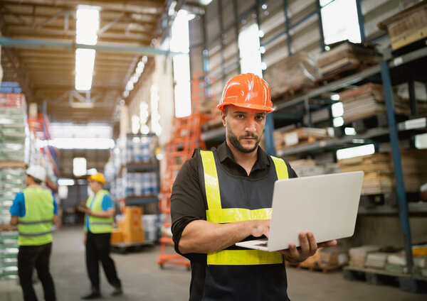Professional manager man employee using laptop check stock working at warehouse. Worker wearing high visibility clothing and a hard hat, helmet and checking and count up goods or boxes for delivery.