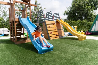 Şirin Asyalı kız gülümsemesi okulda, anaokulunda ya da çocuk parkında sürgülü oynuyor. Çocuklar için sağlıklı bir yaz aktivitesi. Küçük Asyalı kız çocuk parkında dışarı tırmanıyor. Bebek aktivitesi konsepti.