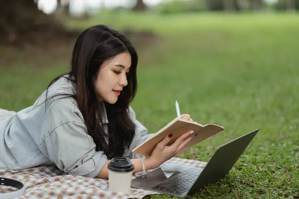 Asian woman smiling lay on picnic blanket and lawn at park garden working on laptop and writing on notebook. Asian female using laptop while sitting under a tree at park. Work from anywhere concept.