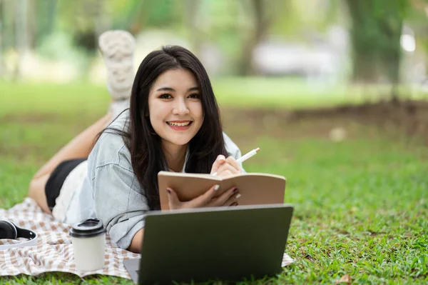 Asian woman smiling lay on picnic blanket and lawn at park garden working on laptop and writing on notebook. Asian female using laptop while sitting under a tree at park. Work from anywhere concept.