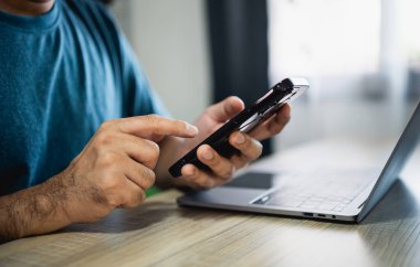 Masada dizüstü bilgisayarla çalıştıktan sonra akıllı telefon kullanan bir adam, iş adamı cep telefonunu arama yapmak veya sosyal medya veya online alışveriş yapmak için kullanıyor. İş konsepti akıllı telefon teknolojisi sohbet konferansı.