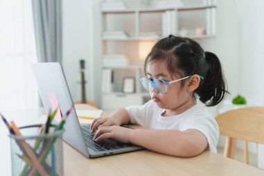 Beyaz tişört ve gözlük giyen Asyalı bir kız bebek evdeki oturma odasındaki ahşap masada dizüstü bilgisayar kullanıyor ve ders çalışıyor. Eğitim öğrenimi online olarak ev kavramından.