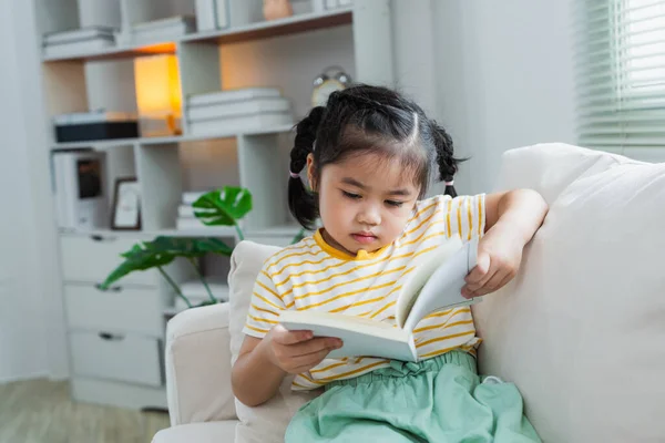 楽しい幸せなアジアの子供のベビーガールは 自宅のリビングルームのソファーに座っている間 笑顔と読書本を読みます 家のソファーで読書本の笑顔をリリース 学校のコンセプトに戻る — ストック写真
