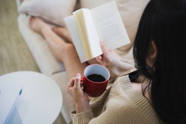 Neşeli Asyalı kadın kitap okuyor ve evdeki oturma odasında kanepede otururken sıcak kahve içiyor. Asyalı kadınlar evdeki kanepede kitap okuyup gülümserler. Ev kavramında etkinlik hobisi.
