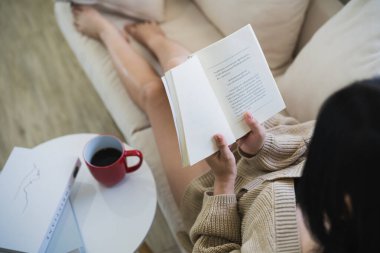 Evdeki oturma odasında kanepede otururken gülümseyen ve kitap okuyan neşeli Asyalı kadın. Asyalı kadınlar evdeki kanepede kitap okuyup gülümsüyor. Ev kavramında etkinlik hobisi.
