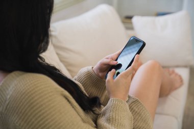 Üst Manzara. Kanepede rahat kıyafetler giyen Asyalı kadın sosyal medyada cep telefonu kullanıyor ya da iş arıyor ya da sohbet ediyor ya da oturma odasında hisse senedi satıyor..