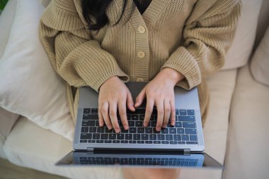 Üst Manzara. Asyalı serbest çalışan kadın gülümseyerek uzanıyor rahatlıyor, klavyede daktilo yazıyor ve dizüstü bilgisayarla kanepenin üzerinde çalışıyor. Girişimci kadın işi için oturma odasında çalışıyor. İş yeri.