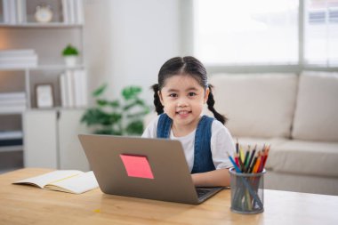 Gülümseyen Asyalı bebek dizüstü bilgisayar kullanıyor ya da evdeki oturma odasındaki ahşap masa üzerinde not defteri çalışmasına resim çiziyor. Eğitim öğrenimi online olarak ev kavramından.