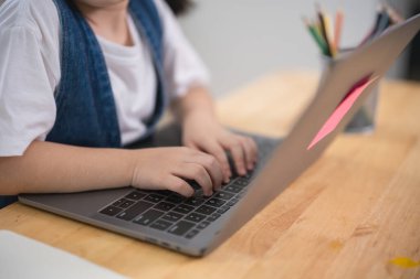 Gülümseyen Asyalı bebek dizüstü bilgisayar kullanıyor ya da evdeki oturma odasındaki ahşap masa üzerinde not defteri çalışmasına resim çiziyor. Eğitim öğrenimi online olarak ev kavramından.