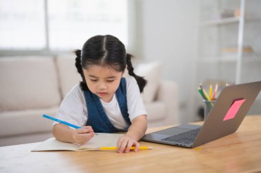 Gülümseyen Asyalı bebek dizüstü bilgisayar kullanıyor ya da evdeki oturma odasındaki ahşap masa üzerinde not defteri çalışmasına resim çiziyor. Eğitim öğrenimi online olarak ev kavramından.