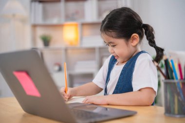 Gülümseyen Asyalı bebek dizüstü bilgisayar kullanıyor ya da evdeki oturma odasındaki ahşap masa üzerinde not defteri çalışmasına resim çiziyor. Eğitim öğrenimi online olarak ev kavramından.