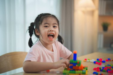 Neşeli Asyalı kız mutlu ve gülümsüyor renkli Lego oyuncakları oynuyor, oturma odasında masanın üzerinde oturuyor, yaratıcı bir şekilde Lego ile oynuyor, renkli yapılar inşa ediyor yaratıcılığı hayal edin..