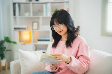 Evdeki oturma odasında kanepede otururken gülümseyen ve kitap okuyan neşeli Asyalı kadın. Asyalı kadınlar evdeki kanepede kitap okuyup gülümsüyor. Ev kavramında etkinlik hobisi.