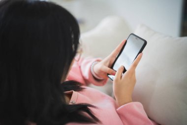 Gülümseyen Asyalı kadın gündelik giysiler içinde kanepede oturuyor sosyal medyada cep telefonu kullanıyor ya da iş arıyor ya da sohbet ediyor ya da oturma odasında hisse senedi satıyor..