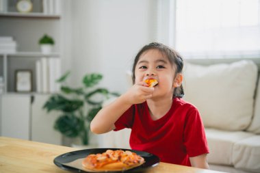 Asyalı mutlu çocuk kız pizza yiyor ve evdeki oturma odasında gülümsüyor. Çocuklar evde İtalyan ev yapımı pizza yiyor ve tadıyor..