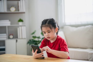 Cep telefonu ekranını kullanan ve dokunan Asyalı bir kız çocuğu. Bebek gülümsemesi cep telefonu kullanmak için komik bir zaman. Çok fazla ekran zamanı. Oturma odasında televizyon seyreden tatlı bir kız, internet bağımlılığı..