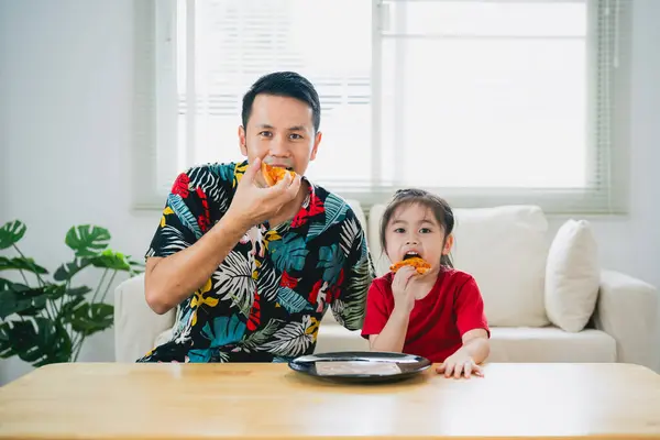 Asian happy children girl and her father feeding eating pizza and smiling in the living room at home. Children girl and her dad eating and tasting italian homemade pizza in house.
